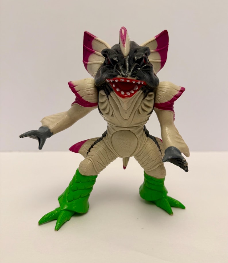 May include: A detailed figurine of a creature with a black head, a wide red mouth, and large, fan-like appendages with pink accents. The figure has tan skin, green legs, and gray hands, showcasing a unique design.