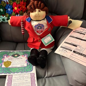 Może przedstawiać: Lalka Cabbage Patch Kids Olympikids z brązowymi włosami z włóczki, ubrana w czerwoną marynarkę, białą koszulkę z logo i czarne buty. Zawiera akt urodzenia i wypis ze szpitala. Lalka trzyma w ręku czerwono-białą pałeczkę sztafetową.