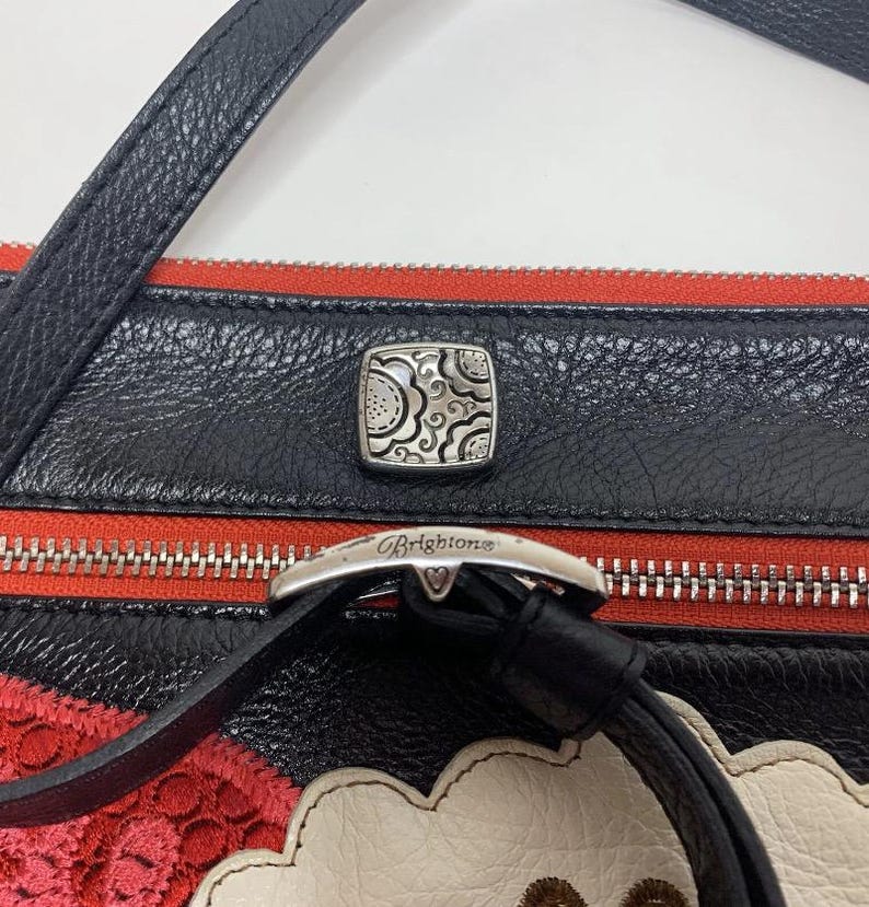 May include: Black leather crossbody bag with red accents and a silver-tone buckle. The bag features a decorative silver square clasp and a black strap. The Brighton brand name is visible on the buckle.