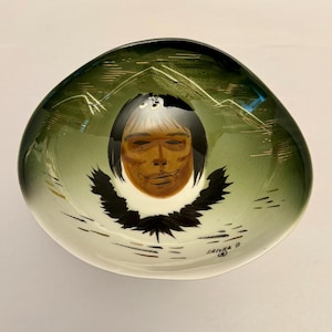 May include: A decorative, oval-shaped bowl with a green and white gradient. The bowl features a painted face with black hair and a black feather design. Mountains are painted around the face. The artist's signature, Jaina B, is visible.