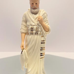 May include: A ceramic figurine of a man in cream-colored robes, holding a white cloth and a shoulder wrap. The figure has a white beard and a turban. Gold accents adorn the sash and robe. The figurine is standing on a reflective surface.