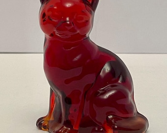 Peut inclure: Figurine de chat en verre rouge rubis, en position assise. Le chat a des oreilles pointues, les yeux fermés et une finition lisse et brillante. La figurine est de couleur unie avec des zones de rouge et d'orange plus claires.