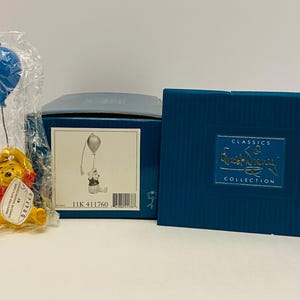 Puede incluir: Una figura de Winnie the Pooh sosteniendo un globo azul, empaquetada en plástico. La figura es amarilla con una camisa roja. Una caja azul con el logotipo de Disney Classics Collection está al lado.