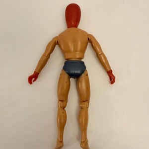 Puede incluir: Una figura articulada de color beige, con cabeza y manos rojas, y calzoncillos azul oscuro. La figura est&aacute; posada con los brazos extendidos y las piernas ligeramente separadas, mostrando su construcci&oacute;n articulada.