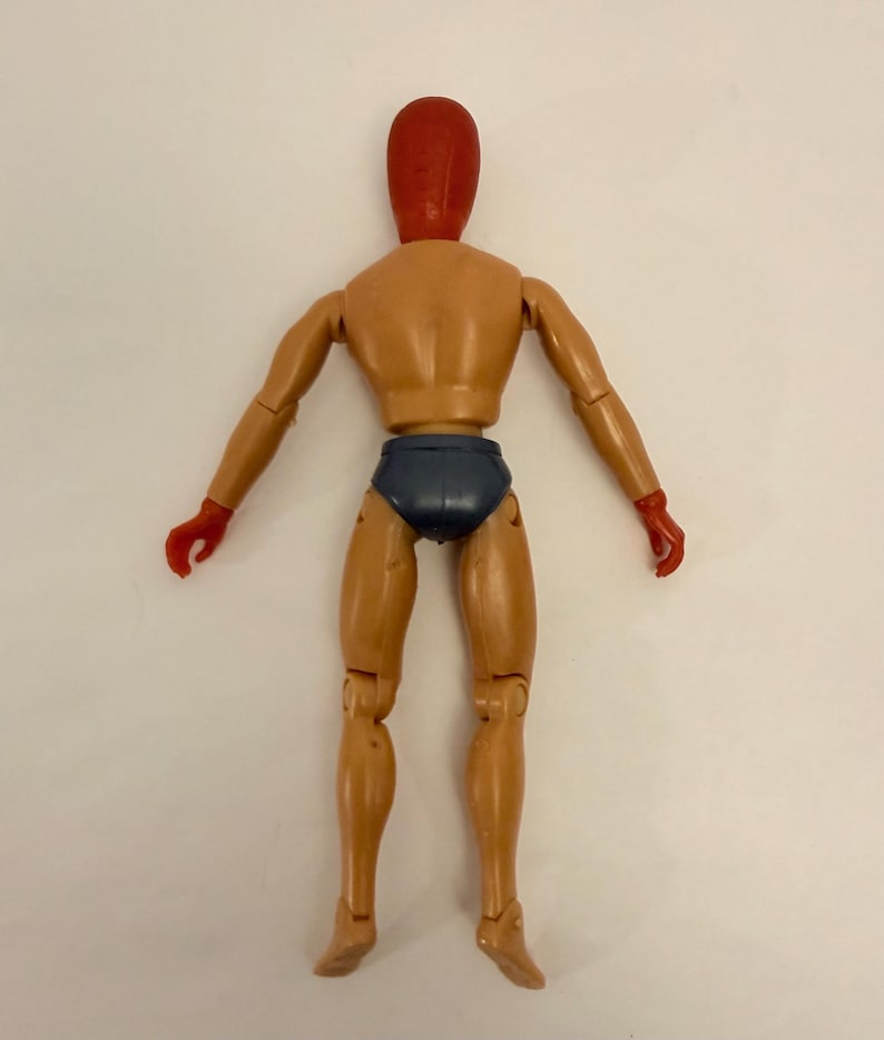 Puede incluir: Una figura articulada de color beige, con cabeza y manos rojas, y calzoncillos azul oscuro. La figura est&aacute; posada con los brazos extendidos y las piernas ligeramente separadas, mostrando su construcci&oacute;n articulada.