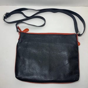 May include: Black leather crossbody bag with a red zipper and trim. The bag has a long, adjustable black strap with a silver buckle. The bag is rectangular and has a textured surface.