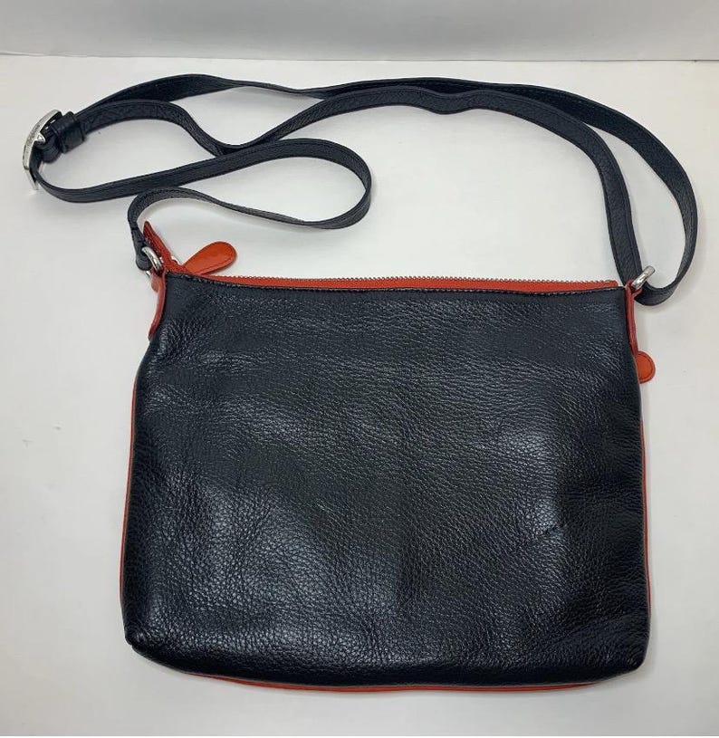May include: Black leather crossbody bag with a red zipper and trim. The bag has a long, adjustable black strap with a silver buckle. The bag is rectangular and has a textured surface.