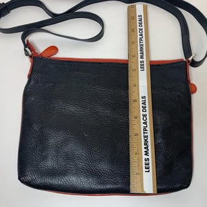 May include: Black leather crossbody bag with a red zipper and trim. The bag has a black adjustable strap and silver hardware. The bag is approximately 10 inches wide.