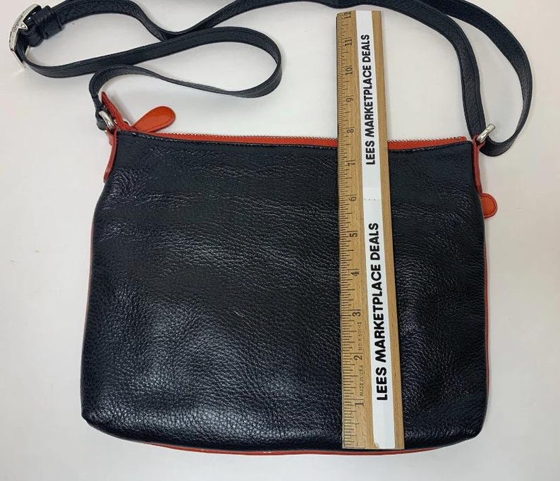 May include: Black leather crossbody bag with a red zipper and trim. The bag has a black adjustable strap and silver hardware. The bag is approximately 10 inches wide.