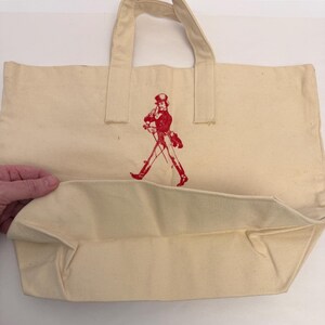 K&ouml;nnte beinhalten: Cremefarbene Canvas-Tragetasche mit roter Grafik eines gehenden Mannes. Die Tasche hat zwei Griffe und eine rechteckige Form. Die Grafik ist eine stilisierte Illustration eines Mannes mit Zylinder und Mantel.