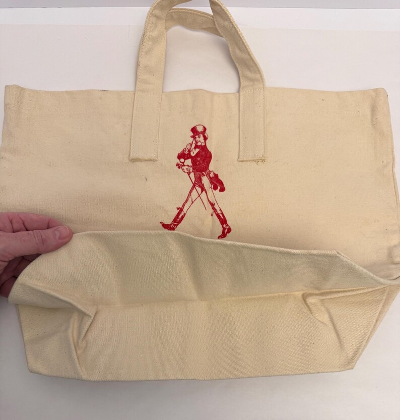 K&ouml;nnte beinhalten: Cremefarbene Canvas-Tragetasche mit roter Grafik eines gehenden Mannes. Die Tasche hat zwei Griffe und eine rechteckige Form. Die Grafik ist eine stilisierte Illustration eines Mannes mit Zylinder und Mantel.