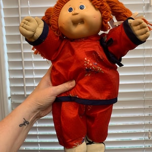 Może przedstawiać: Lalka Cabbage Patch Kid z czerwonymi włosami ubrana w chiński strój w kolorze czerwonym i niebieskim z kwiatowymi haftami. Lalka ma niebieskie oczy i szeroki uśmiech z zębami.