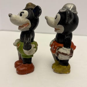 May include: Two vintage Mickey Mouse figurines. One Mickey figurine wears a brown hat, green shorts, and red shoes. The other Mickey figurine wears a silver hat, red skirt with white polka dots, and yellow shoes. Both figurines have black bodies and white faces.