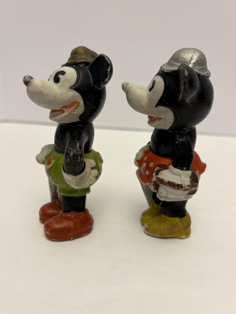 May include: Two vintage Mickey Mouse figurines. One Mickey figurine wears a brown hat, green shorts, and red shoes. The other Mickey figurine wears a silver hat, red skirt with white polka dots, and yellow shoes. Both figurines have black bodies and white faces.