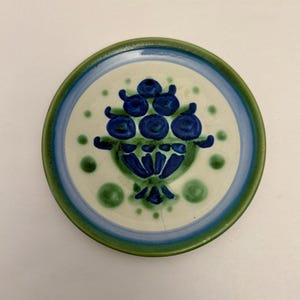 Könnte beinhalten: Runder Keramikteller mit handgemaltem Design. Der Teller hat ein zentrales Motiv aus blauen floralen Elementen innerhalb eines grünen und blauen Randes. Grüne und blaue Punkte sind um das florale Design verstreut.