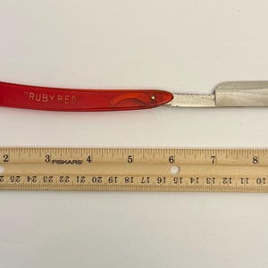 Puede incluir: Una navaja de afeitar con mango rojo, marcada "RUBY RED", y una hoja plateada, junto a una regla de madera. La navaja mide aproximadamente 25 cm de largo, ideal para afeitarse y acicalarse.