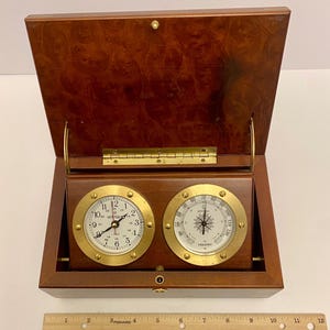 May include: A wooden box with a hinged lid, housing a clock and a thermometer. The clock face is white with black numerals and hands, and the thermometer has a compass rose design. The box is approximately 10 inches wide.