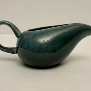 May include: A teal-colored ceramic gravy boat with a rounded body, a curved handle, and a spout. The gravy boat has a glossy finish and sits on a white surface. The handle is a circular shape.