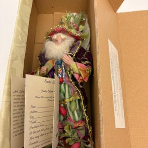 May include: A detailed figurine of a fairy or gnome, dressed in a colorful outfit with a purple velvet coat, striped pants, and a decorative hat. The figurine is inside a cardboard box, with a tag that says "Easter B" and "Limited Edition".