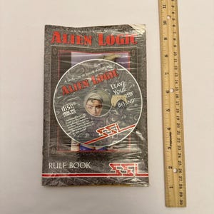 May include: A sealed Alien Logic game package with a CD-ROM and rule book. The CD-ROM features the game title and a portrait of a person. The rule book is visible beneath the CD. A wooden ruler is placed next to the game package.
