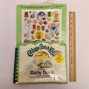Op de afbeelding: Een Cabbage Patch Kids stickerboekset. De set bevat een stickervel met diverse karakterstickers en een stickerboek van 16 pagina's. De kaft van het boek bevat de tekst "Cabbage Patch Kids Sticker Baby Book". Een liniaal staat naast het boek.