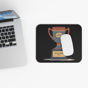 May include: A black mousepad with a trophy design. The trophy is blue, brown, and gold, with the text "Best Father Ever" and space for a name. A white mouse is next to the mousepad.