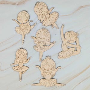 May include: Six wooden ballerina cutouts in various poses. Each cutout features a ballerina with a crown, tutu, and ballet shoes. The cutouts are made of light-colored wood and are arranged on a light-colored surface.
