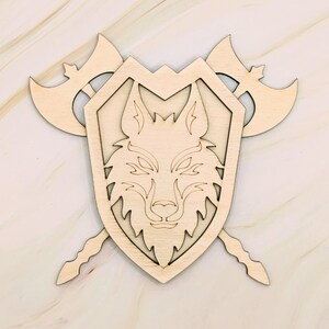 May include: A wooden shield with a wolf's head design, flanked by two axes. The shield and axes are made of light-colored wood, with the wolf's head carved into the center of the shield. The axes are positioned behind the shield.