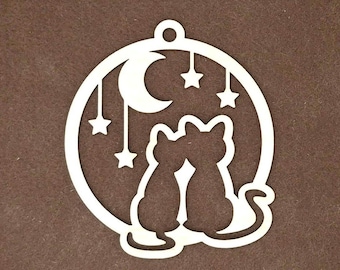 Op de afbeelding: Een cirkelvormig ornament van lichtgekleurd hout met een uitgesneden ontwerp van twee katten die naast elkaar zitten. Boven de katten zijn een maansikkel en drie hangende sterren afgebeeld binnen het ronde frame, dat aan de bovenkant een klein gaatje heeft.