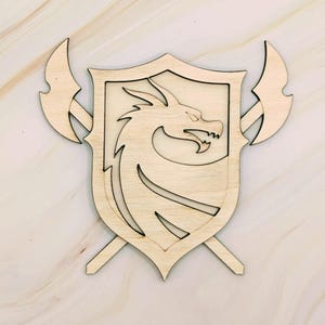 May include: A wooden shield with a dragon emblem and two crossed axes. The shield is light brown with a dragon silhouette in the center. The axes are positioned behind the shield, creating a medieval or fantasy theme.
