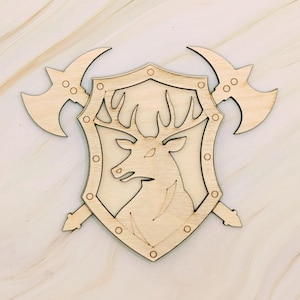 May include: Wooden shield with a deer head emblem, flanked by two axes. The shield has a decorative border with rivets. The axes are positioned behind the shield, creating a heraldic design. The wood has a natural, light color.