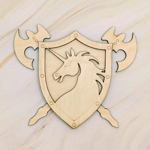 May include: Wooden shield with a unicorn design, crossed with two axes. The shield is light brown with a detailed unicorn silhouette. The axes are also light brown and are positioned behind the shield. The design is likely for a wall hanging or decorative item.