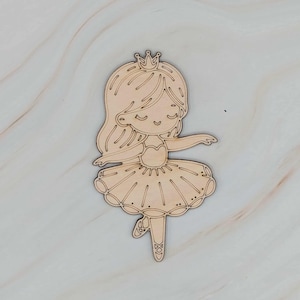 May include: A wooden cutout of a ballerina with a crown. The ballerina has long hair, a tutu, and ballet slippers. The cutout is made of light-colored wood and has a simple, cartoon-like design. The ballerina is in a dancing pose.