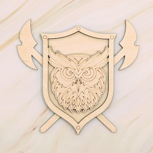 May include: Wooden wall art featuring a shield design with an owl in the center, flanked by two axes. The owl has detailed feather and eye markings. The shield has decorative rivets. The art is made of light-colored wood.