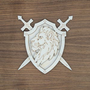 May include: A light-colored wooden cutout featuring a shield emblem with a lion head profile in the center. Two crossed swords are positioned behind the shield, with their hilts extending from the top and blades pointing downward at the sides.