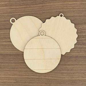 May include: Three unfinished wooden Christmas ornaments in various shapes: a circle, a scalloped edge shape, and a round ornament with horizontal lines. Each ornament has a small hole for hanging. The ornaments are light brown.