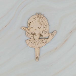 May include: A wooden cutout of a ballerina in a tutu and crown. The ballerina is in a graceful pose with one leg extended. The cutout is made of light-colored wood, with etched details for the face, hair, and tutu.