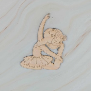 May include: A wooden cutout of a ballerina in a seated pose, with one arm raised and the other bent. The ballerina wears a tutu. The wood has a light, natural color. The cutout is on a light-colored, marbled surface.
