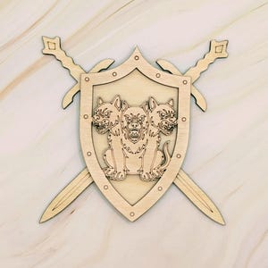 May include: Wooden shield with a three-headed dog emblem, flanked by two crossed swords. The shield has a light brown finish with a detailed carving of the dog. The swords have decorative hilt designs.