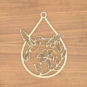 May include: A light-colored wooden cutout ornament in a teardrop shape featuring a laser-cut design of a hummingbird hovering near two lily flowers. The ornament includes a small circular hole at the top for hanging, with a smooth finish.