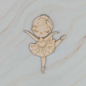 May include: A wooden cutout of a ballerina in a tutu and crown. The ballerina is in a graceful pose, with one leg extended and arms outstretched. The cutout is made of light-colored wood and has a smooth finish.