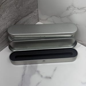 May include: Three silver-colored metal pen cases, one open to reveal a black interior. The cases are rectangular with rounded edges. The open case has a black foam insert. The cases are on a white and gray marble-patterned surface.