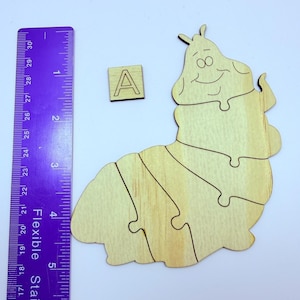 May include: A wooden caterpillar puzzle with a smiling face, composed of several interlocking pieces. A small wooden square with the letter "A" is next to the puzzle. A purple ruler is placed next to the puzzle, with measurements in inches.