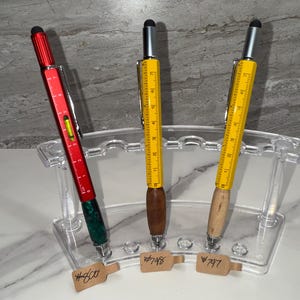 Peut inclure: Trois stylos multifonctions dans un support en acrylique transparent. L'un est rouge, les autres jaunes avec des marquages de règle. Les stylos ont des accents en bois et en métal. Chaque stylo a une pointe stylet.
