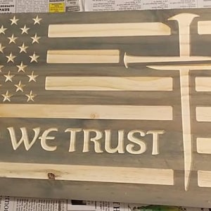 May include: A rectangular wooden wall sign featuring a carved American flag design with a gray-stained finish. The piece includes carved stars, horizontal stripes, a cross formed by two nails, and the text IN GOD WE TRUST in large capital letters.