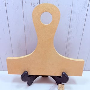 May include: A light tan, flat, wooden cutout shape featuring a wide rectangular base and a tall, narrow neck with a circular hole at the top. The object is supported by a black display stand and includes a small square tile marked with the letter A.