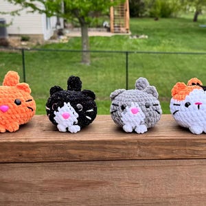 May include: Four crocheted cat-shaped toys in a row. The cats are orange, black, gray, and calico, each with a pink nose and black whiskers. They are sitting on a wooden surface, with a green background.