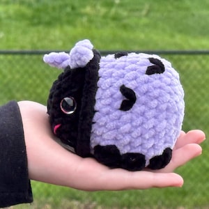 May include: A handmade crochet plushie in the shape of a cow. The plushie is black and lavender with black spots. It has small lavender horns and a pink mouth. The plushie is held in a person's hand.
