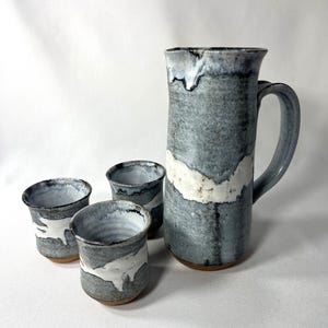 May include: A handmade ceramic pitcher and three matching cups. The pitcher and cups are glazed in shades of gray and white, with a textured appearance. The pitcher has a handle and a wide spout. The cups are small and cylindrical.