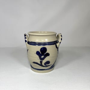 May include: A small, off-white ceramic vase with two handles and a dark blue floral design. The vase features two dark blue horizontal stripes near the top and bottom. The floral design is centered on the front of the vase.